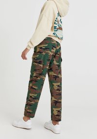 Groene, bruine en zwarte camouflagedraagbroek met meerdere zakken en een relaxte pasvorm. Gecombineerd met een crèmekleurige hoodie.