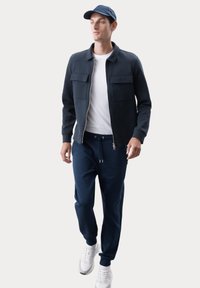 Giacca bomber navy con due tasche frontali, indossata sopra una t-shirt bianca. Abbinata a joggers navy e sneakers bianche, completa un look casual.
