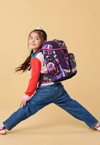 Sac à dos avec une base violette, des motifs colorés, des accents roses et un logo circulaire. Un enfant le porte, posant sur un fond beige.