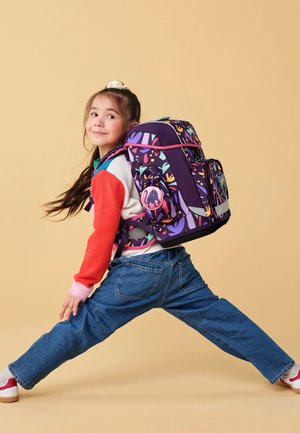 Rucksack mit lila Grundfarbe, bunten Mustern, rosa Akzenten und einem runden Logo. Ein Kind trägt ihn und posiert vor einem beigen Hintergrund.