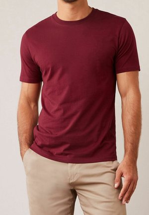 Man wearing a plain burgundy short-sleeve T-shirt and beige pants, with one hand in pocket and neutral background.