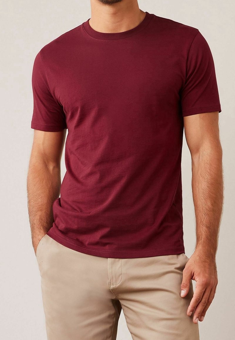 Homme portant un T-shirt bordeaux uni à manches courtes et un pantalon beige, une main dans la poche et un fond neutre.