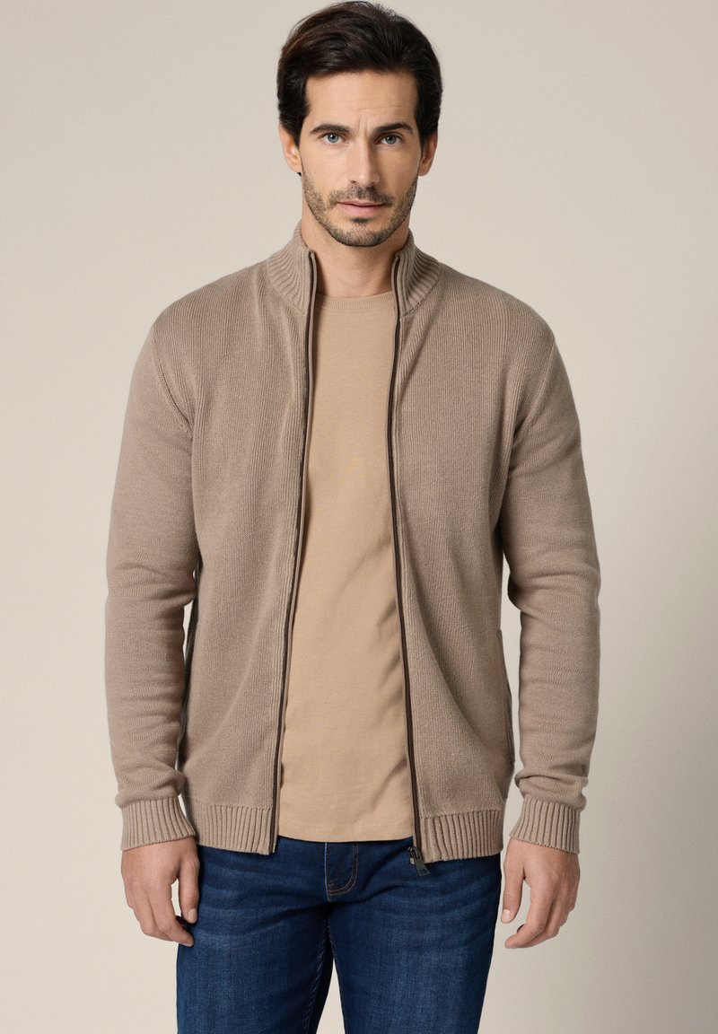 Mężczyzna ubrany w beżowy sweter rozpinany na zamek, nałożony na jasnobrązową koszulę i niebieskie dżinsy, stojący na tle jednolitego jasnego tła.