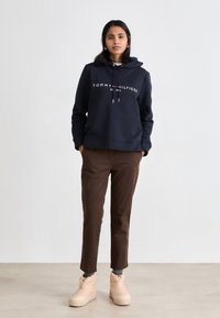 Marine hoodie met witte logo-tekst, bruine taps toelopende broeken en beige robuuste laarzen. De materialen lijken zacht; de hoodie heeft een trekkoordfunctie.