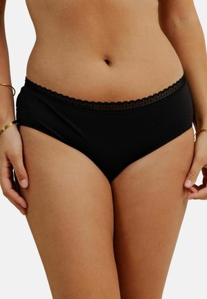 Femme portant un slip en coton noir avec une bordure en dentelle à la taille, se tenant avec les mains détendues sur les hanches, sur un fond blanc uni.