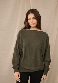 Noor naine pikkade pruunide juustega, seljas oliivrohelise pika varrukaga kampsun ja mustad püksid, seismas neutraalse kangataustaga.