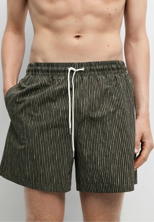 Persona a torso nudo che indossa pantaloncini a righe verticali nere e verde oliva con coulisse bianca, mani in tasca, in piedi su uno sfondo semplice.