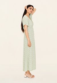 Hellgrünes, florales Maxikleid mit kurzen Ärmeln, das mit gelben und weißen Mustern verziert ist. Kombiniert mit pinken, gestrickten Sandalen.