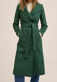 Grøn uldblandingsfrakke med reverskrave, bælte i taljen og knapper foran. Har lange ærmer og sidelommer. Glat tekstur.