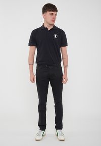 Polo shirt nera a maniche corte, con logo sul petto, abbinata a pantaloni scuri e sneakers bianche con accenti verdi.