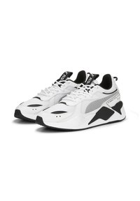 Des baskets blanches avec des accents noirs, présentant un design à lacets, en matériau synthétique et en mesh, avec une semelle texturée et un motif rayé sur le côté.