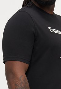 Must musta T-särk, mis on valmistatud venivast kangast, valgete tikandite tekstiga rinnal ja tugevdusõmblustega õmblustes.