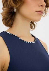 Abito blu navy con scollatura rotonda decorata con piccoli strass trasparenti. Il tessuto appare liscio e aderente. Capelli testurizzati visibili.