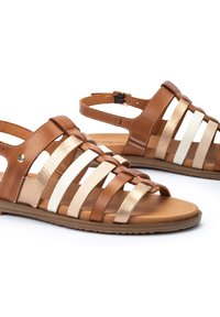 Sandalias de cuero marrón que presentan múltiples correas entrelazadas en tonos claros y oscuros con acentos metálicos, suela plana y correa ajustable en el tobillo.