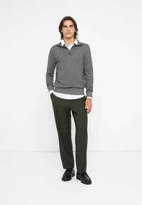 Maglione grigio con zip e colletto, abbinato a pantaloni verdi oliva. Il modello indossa scarpe nere. Tessuto semplicemente strutturato e linee pulite.