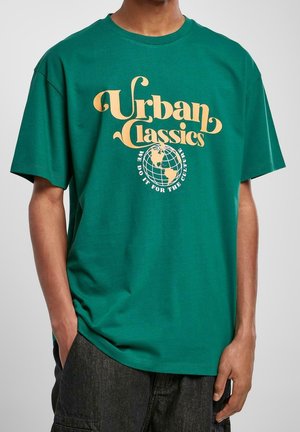 Grünes T-Shirt mit gelbem Text "Urban Classics" über einer Grafik einer Weltkugel und dem Spruch "We do it for the culture", getragen von einer Person mit Händen in den Taschen.