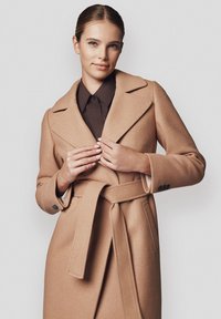 Jasnobrązowy wełniany płaszcz z podwójnym zapięciem, szerokimi klapami, paskiem w talii i zapięciami na guzik. Nosi się go na ciemnobrązowej koszuli.