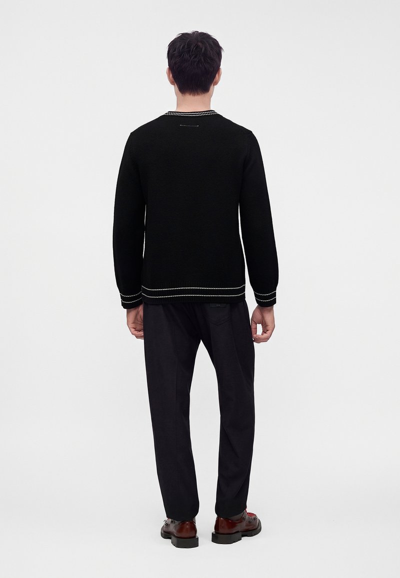 Uomo in piedi di spalle, indossa un maglione nero con bordi bianchi, pantaloni neri e scarpe marrone scuro su uno sfondo chiaro e uniforme.