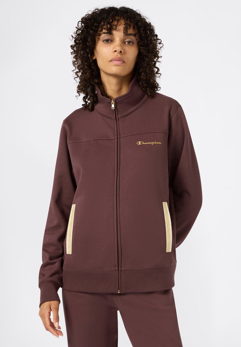 Bordeaux zip-up jack met gouden logo en beige zijzakken, voorzien van een hoge kraag en gestructureerde stof. Lange mouwen met geribde manchetten.
