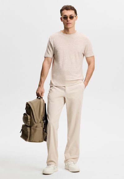 Homme portant un t-shirt beige, un pantalon crème, des baskets blanches, des lunettes de soleil, tenant un sac à dos kaki dans une main sur un fond uni.
