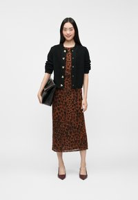 Črna pletena jopica z gumbki v obliki srca, prekrita čez rjav leopardji print obleko. Model drži črno torbico in nosi bordo čevlje s peto.