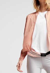 Mujer con una chaqueta rosa claro con cremallera, camiseta blanca de Nike, leggins grises claros y una bolsa de cintura rosa con rayas reflectantes.