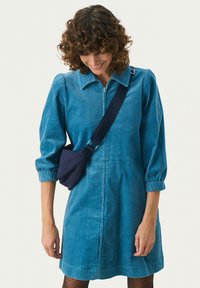 Il vestito in corduroy blu presenta una chiusura con zip frontale, maniche lunghe a sbuffo e una vestibilità morbida. Una borsa a tracolla blu navy aggiunge un tocco funzionale.