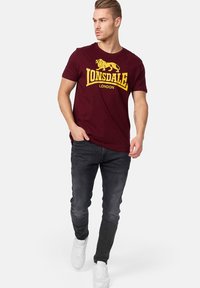Burgundi pamut póló sárga Lonsdale logóval és oroszlán grafikával, sötét farmernadrággal és fehér sportcipővel párosítva. Modell előre sétál.