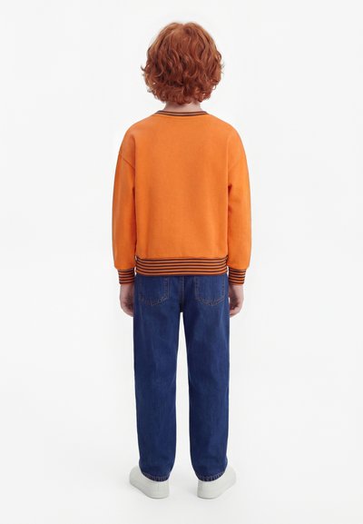 Niño con cabello rizado rojo visto de espaldas, lleva una sudadera naranja con puños a rayas, jeans azules y zapatillas blancas.