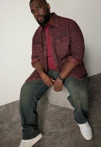 Rustinvärgad denimskjorta över en vinröd t-shirt, kombinerad med mörka denimjeans och vita sneakers. Tillbehören inkluderar armband på handleden.