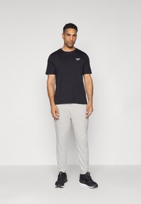T-shirt en coton noir avec un petit logo blanc, associé à un pantalon de survêtement gris clair. Le modèle porte des chaussures de sport noires. Design et coupe simples.