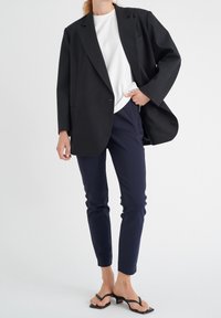Černý oversized blazer, bílá tričková košile s kulatým výstřihem, navy slim-fit kalhoty a černé sandály na pásku. Hladké textury, čisté linie, moderní vzhled.