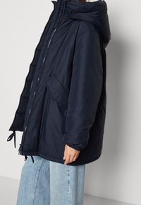 Giacca puffer navy con cappuccio, dotata di polsini elasticizzati, chiusura con zip e tasche laterali. Tessuto liscio e vestibilità oversize.