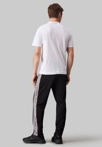 T-shirt bianco a maniche corte con orlo dritto, abbinato a pantaloni neri con strisce laterali grigio chiaro e un logo. Scarpe da ginnastica nere.