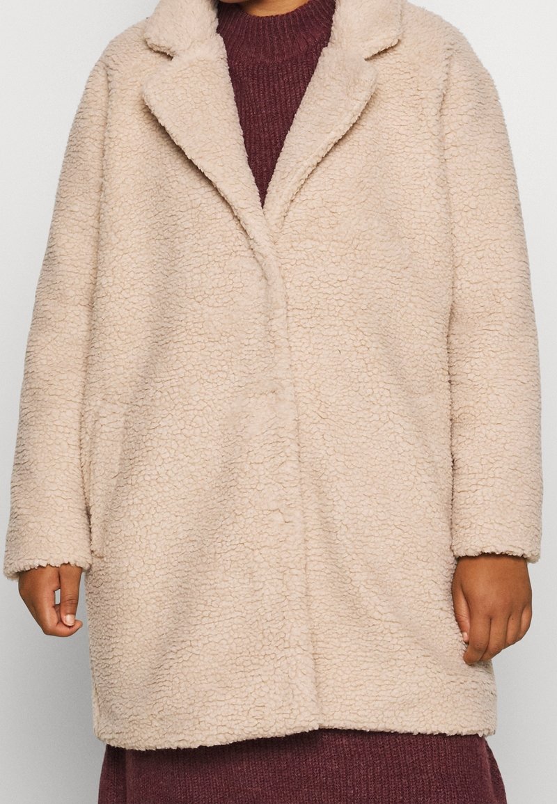 Beige fuskpälsjacka med en strukturerad yta, slagkrage och en avslappnad passform, med långa ärmar och en enkel knappstängning.