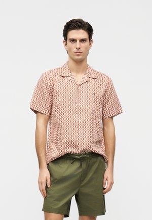Young man wearing a short-sleeve orange patterned shirt and olive green drawstring shorts standing against a white background.