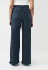 Mørkeblå vide jeans med høj talje, der har to baglommer og en ren søm. Stoffet har en glat, denimtekstur.