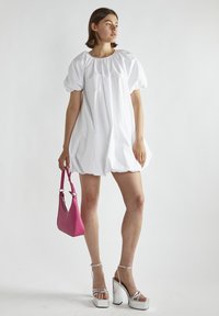 Hvid oversized kjole med pufærmer, elastisk halsudskæring, parret med en pink håndtaske og sølvfarvede plateauhæle. Glat tekstur og voluminøs form.