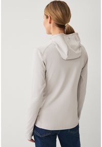 Mujer con coleta rubia vistiendo chaqueta con capucha gris claro y vaqueros azules, de espaldas sobre un fondo liso.