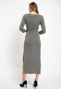 Langärmliges, figurnahes Kleid aus grauem, strukturiertem Stoff, mit einem dezenten Schlitz hinten und einem kleinen dekorativen Akzent am Halsauschnitt. Schwarze Highheels.