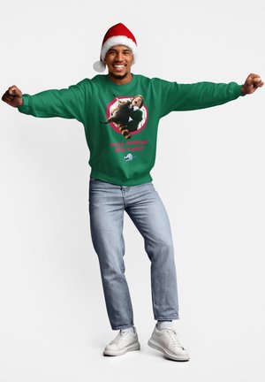 Grøn sweatshirt med grafisk tryk og teksten "Har nogen brug for et kram?" Modellen bærer en rød nissehat, lyseblå jeans og hvide sneakers.