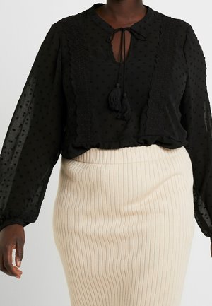 Zwarte doorschijnende blouse met getextureerde polka dots, een geschulpte rand en touwtjes met kwastjes, gecombineerd met een crèmekleurige geribbelde rok.