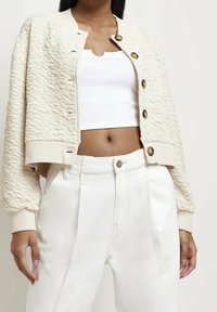 Teksturoitu kerma bomber-takki suurilla napilla, ribbed hihansuilla ja lyhyellä leikkauksella, yhdistettynä korkeavyötäröisiin valkoisiin housuihin.