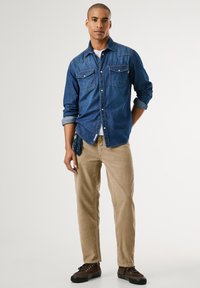Camicia di jeans con due tasche sul petto, maniche arrotolate; abbinata a pantaloni beige a gamba stretta e sneakers marroni. Texture liscia, design casual.