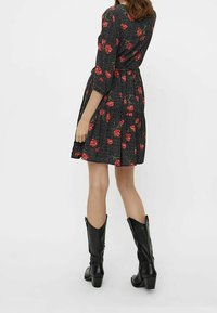 Robe noire à motif floral rouge et pois blancs, ceinturée à la taille, portée avec des bottes noires montantes.