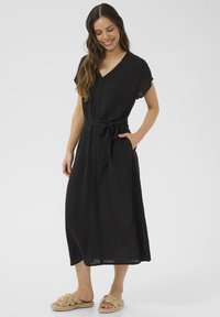 Svart lin klänning med v-ringning, korta ärmar, ett knytbälte i midjan och sidofickor, tillsammans med beige flätade sandaler.