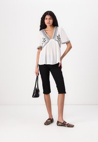 Hvid bluse med sort broderi, V-hals og korte pufærmer. Sammen med sorte capri-bukser og læderneprint ballerinaer. Sort håndtaske.
