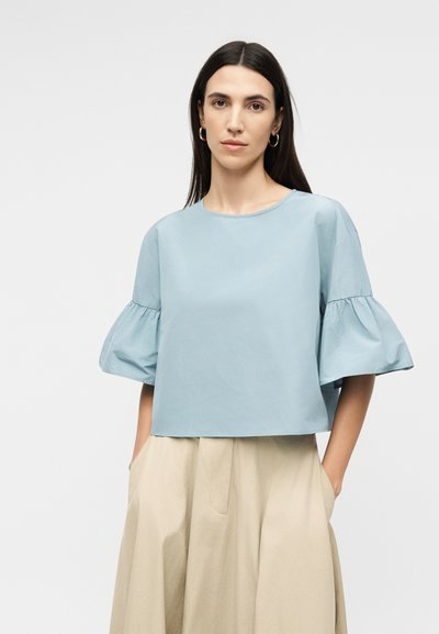 Γυναίκα με μακριά σκοτεινά μαλλιά φοράει ανοιχτό μπλε μπλούζα με κοντά μανίκια και φουσκωτές μανσέτες, καθώς και μπεζ παντελόνι ψηλόμεσο, φαρδύ στο κάτω μέρος, στέκεται με τα χέρια στις τσέπες.