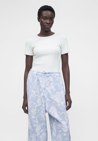 Γυναίκα που φοράει απλό λευκό τοπ με κοντά μανίκια και ανοιχτό μπλε παντελόνι με φαρδιά πόδια, λευκά ανθισμένα σχέδια και κορδόνι στη μέση.