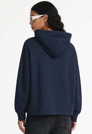 Femme aux longs cheveux noirs portant des lunettes de soleil blanches, un sweat à capuche bleu marine et un jean noir, de dos sur un fond blanc.